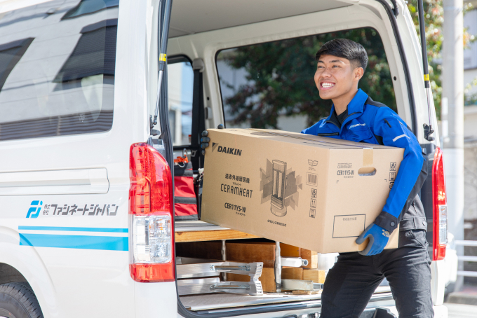 作業着姿の男性が、屋外で白いバン（株式会社ファシネートジャパンの記載あり）からダイキン製機器の段ボール箱を笑顔で運んでいる様