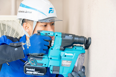 ヘルメットと作業着を着用した男性が、屋外の壁面に対してマキタ製の電動ドリルを使って作業をしている様子